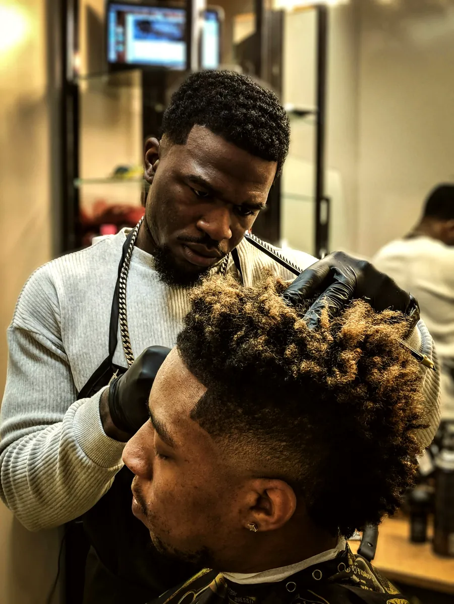 A barber shaping a curly high-top with clippers in the shop.